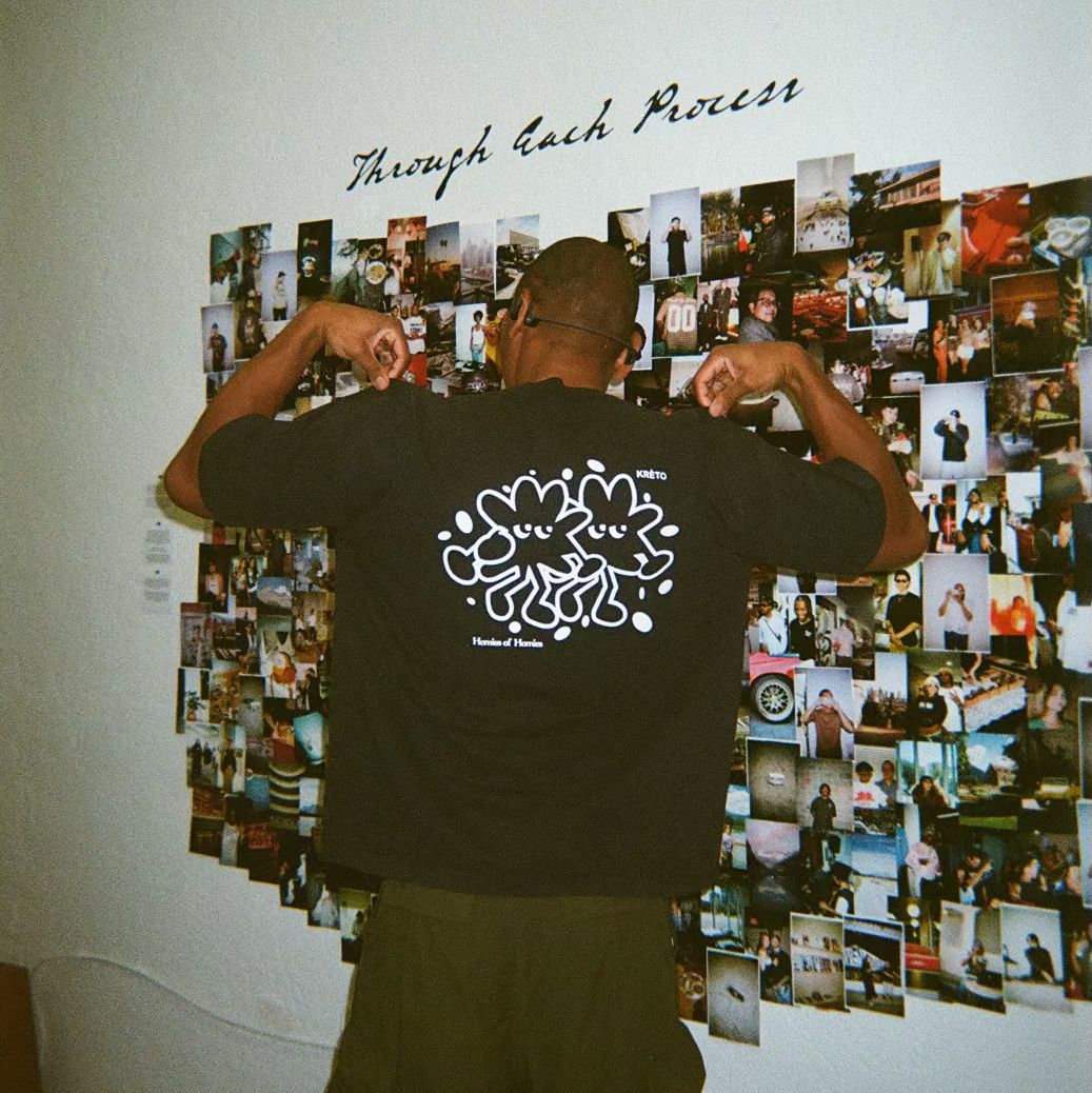 A man stands with his back to the camera, flexing his biceps while wearing a black graphic t-shirt and dark pants. The back of the shirt features a large white abstract graphic and the text 'Through each process'.